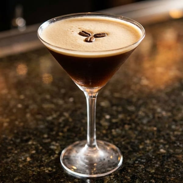 Espresso martini with three coffee beans on top, sitting on a dark, speckled bar.
